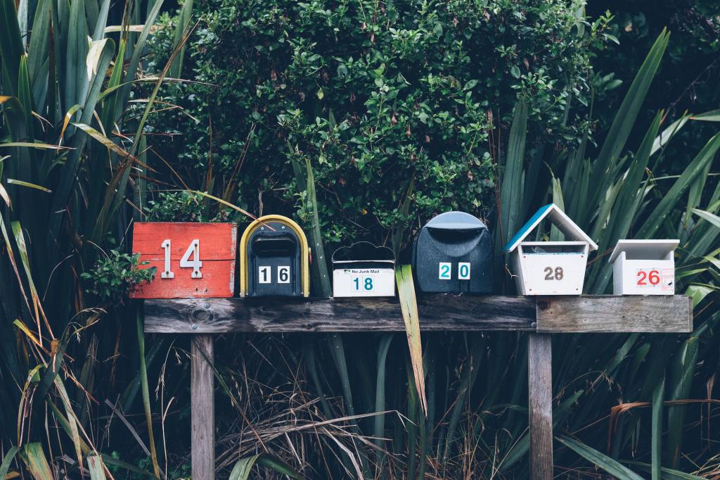 six assorted-color mailboxes in a wooden shelf in front of bushes